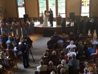 A wedding ceremony in a spacious room, with guests seated in rows of chairs, surrounded by floral decorations.